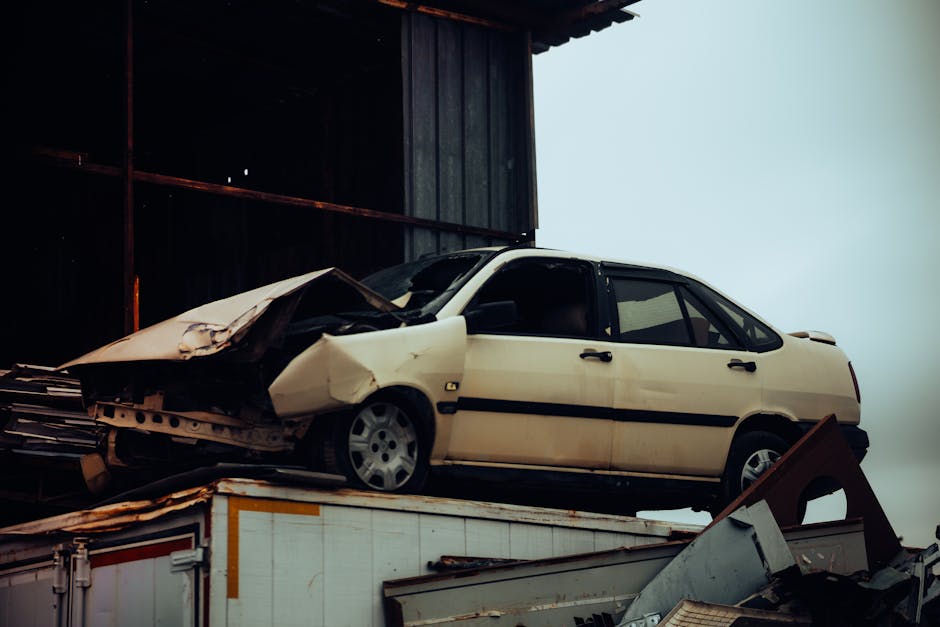 An abandoned white sedan with significant front-end damage, including a crumpled hood and missing front bumper, is positioned on top of a white trucks or container platform. The car's exterior has pat