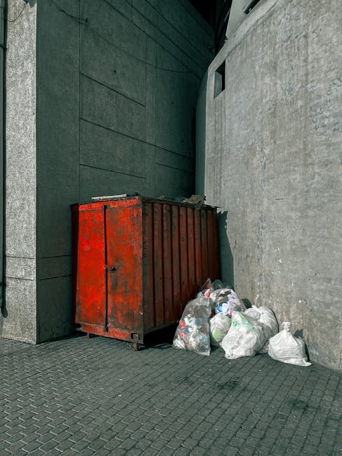 A collection of overflowing rubbish bins and scattered waste on a paved sidewalk adjacent to a parking area. The central large gray bin marked for mixed paper and card is filled to capacity with paper